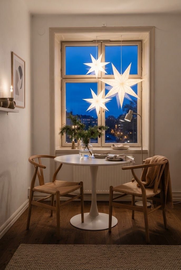 White paper star lanterns glowing in a window above a small round dining table with wishbone chairs at twilight